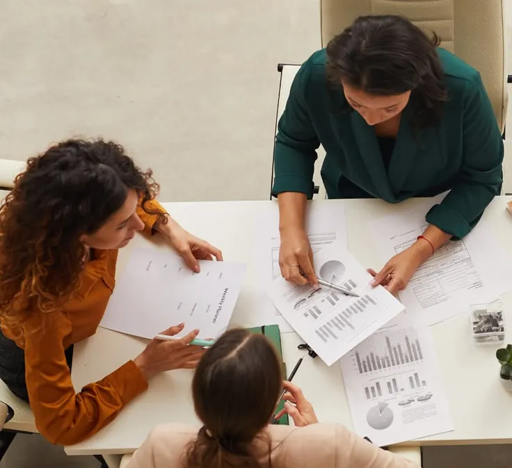 Three colleagues around a table covered in charts and reports, seen from above